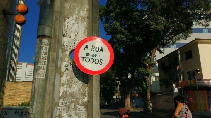 Placa de trânsito presa em um poste, arredondada com contorno vermelho e centro branco, com a frase "A rua é de todos" escrita em letras pretas.